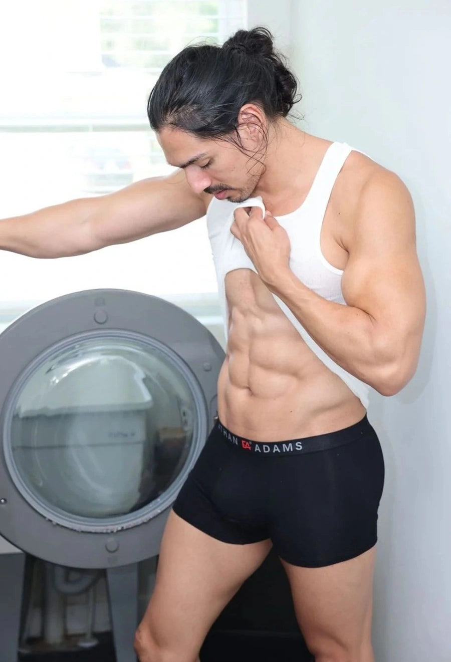 Person wearing a white tank top and black underwear with 'Adams' branding, standing in front of a washing machine. shop confidence Ethan Adams